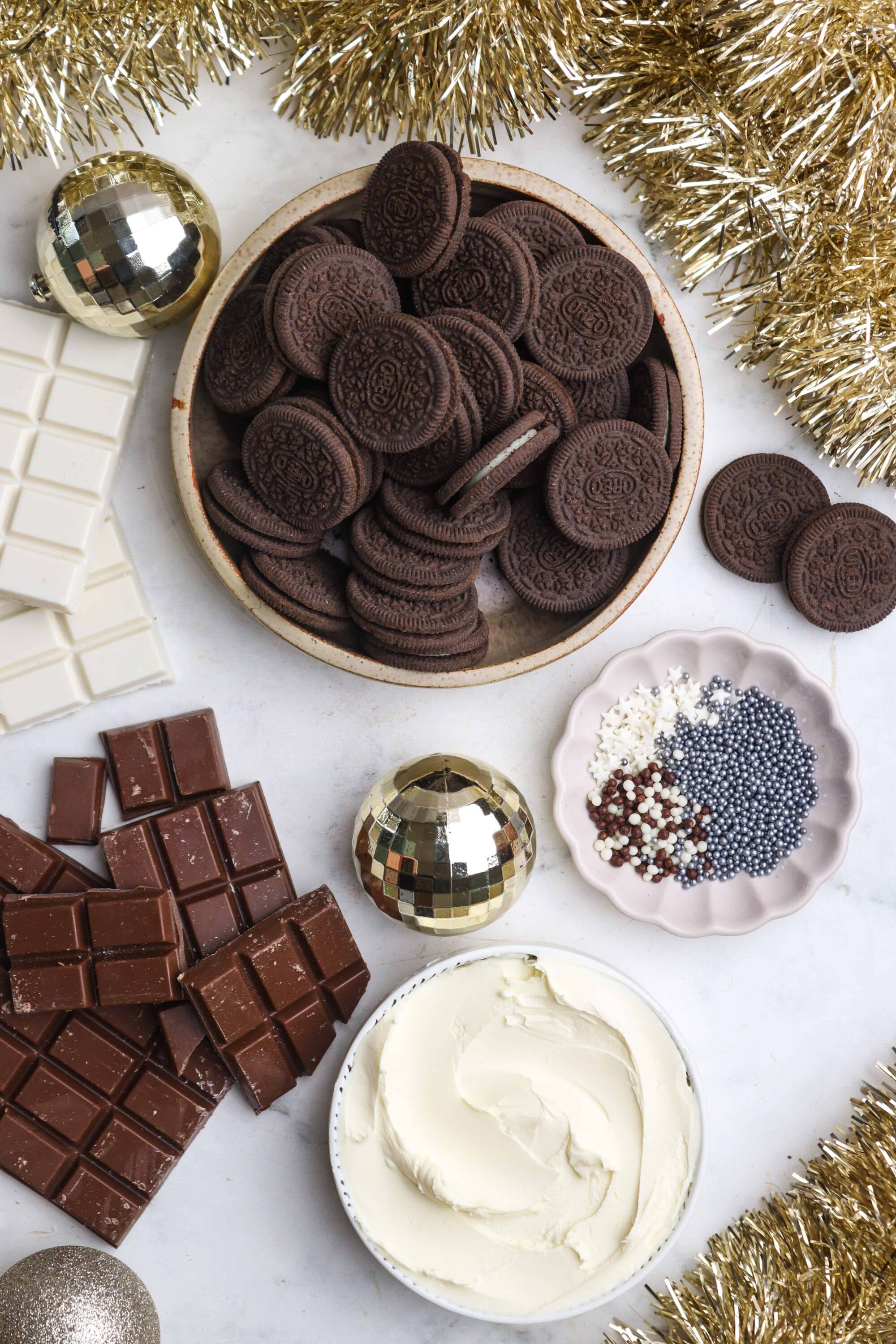 Chocolate Oreo cookies with white chocolate bars, sprinkles, and festive gold and silver ornaments for holiday baking.