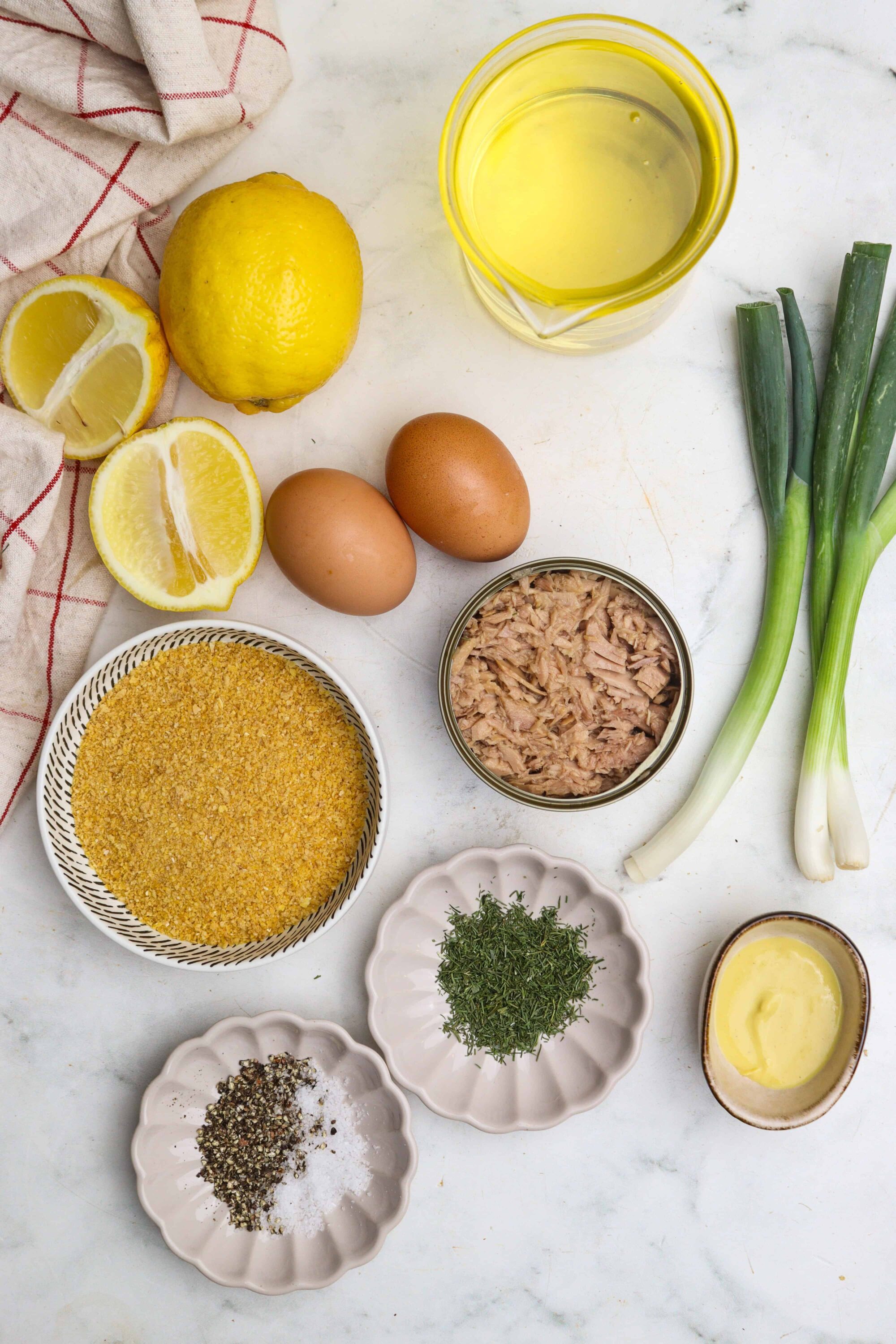 Lemon, eggs, tuna, green onions, dill, breadcrumbs, olive oil, and spices on a white marble surface for tuna salad.