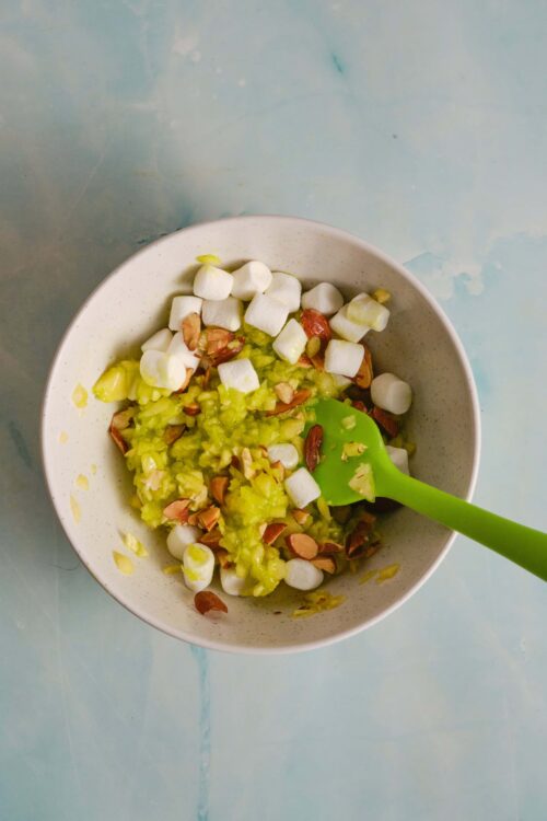 Creamy avocado and toasted almond salad with mini marshmallows in a white bowl, on a blue surface.