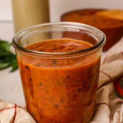 Chunky marinara sauce in a glass jar with basil and kitchen background.