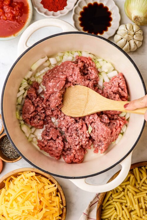 Minced beef browning in a skillet with chopped onions, surrounded by pasta, cheese, and seasonings.