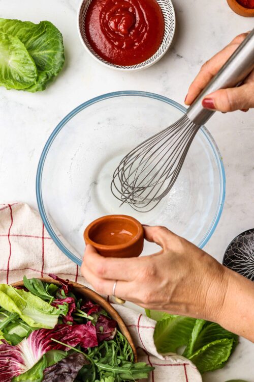 Whisking eggs for homemade salad dressing with fresh greens and tomatoes on a white kitchen countertop.