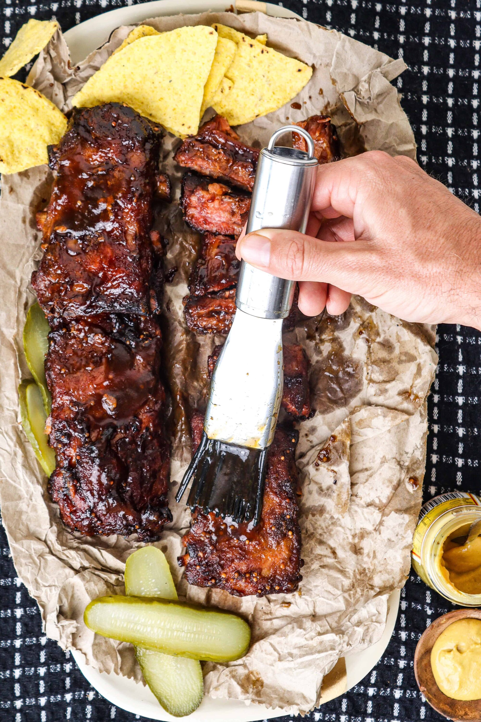 Succulent barbecue ribs glazed with sauce, served with pickles, corn chips, and mustard on parchment paper.