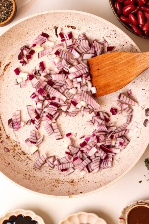 Diced red onions in a beige skillet with a wooden spatula ready for cooking.