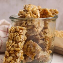 Crispy peanut brittle bars in jar with broken pieces scattered around.