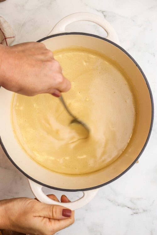 Creamy custard eggs in a pot, being stirred on a white marble countertop.