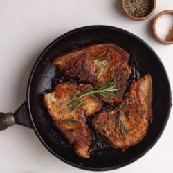 Braised pork chops in a skillet with herbs and seasoning, served with salt and pepper, on a white kitchen countertop.