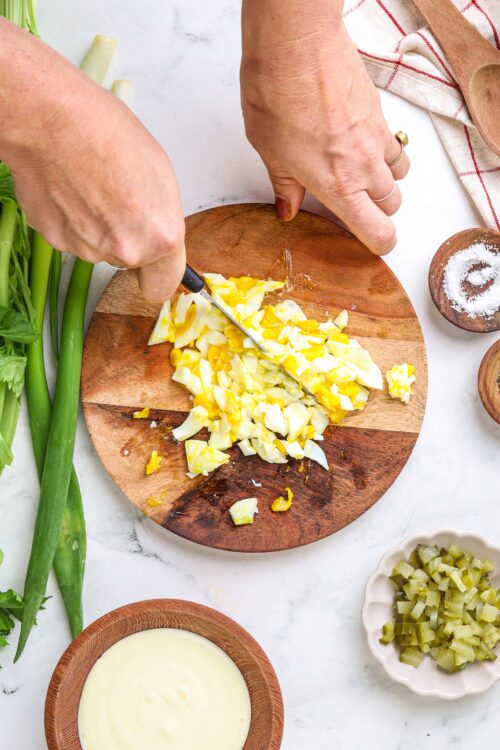 Chopped hard-boiled eggs on a wooden cutting board with fresh green onions and kitchen utensils, perfect for egg salad recipes.