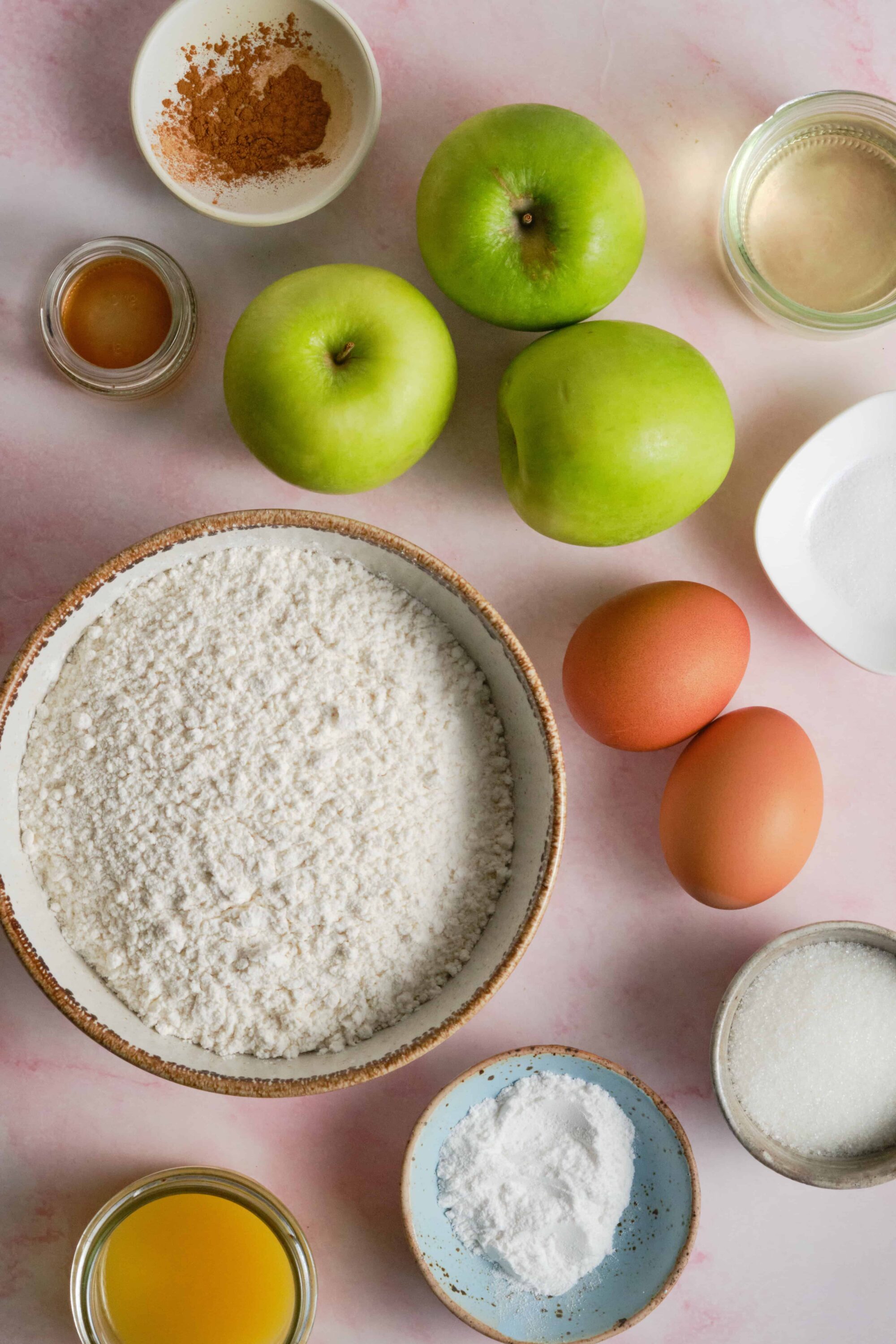 Fresh ingredients for baking apple cinnamon cake, including green apples, eggs, flour, spices, and liquids.