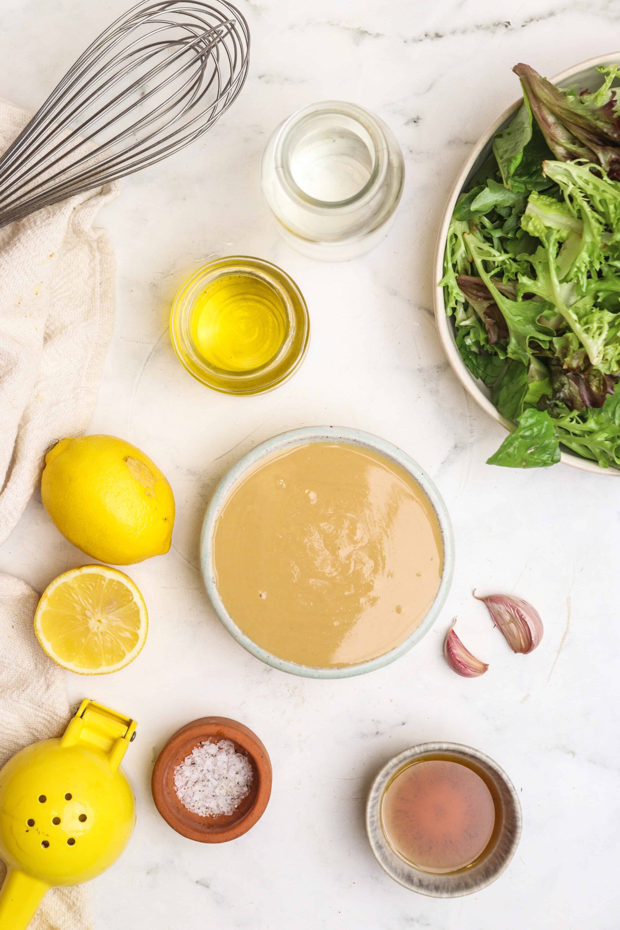 Vinaigrette salad dressing ingredients on white marble surface, olive oil, garlic, lemon, salt, vinegar, fresh greens.