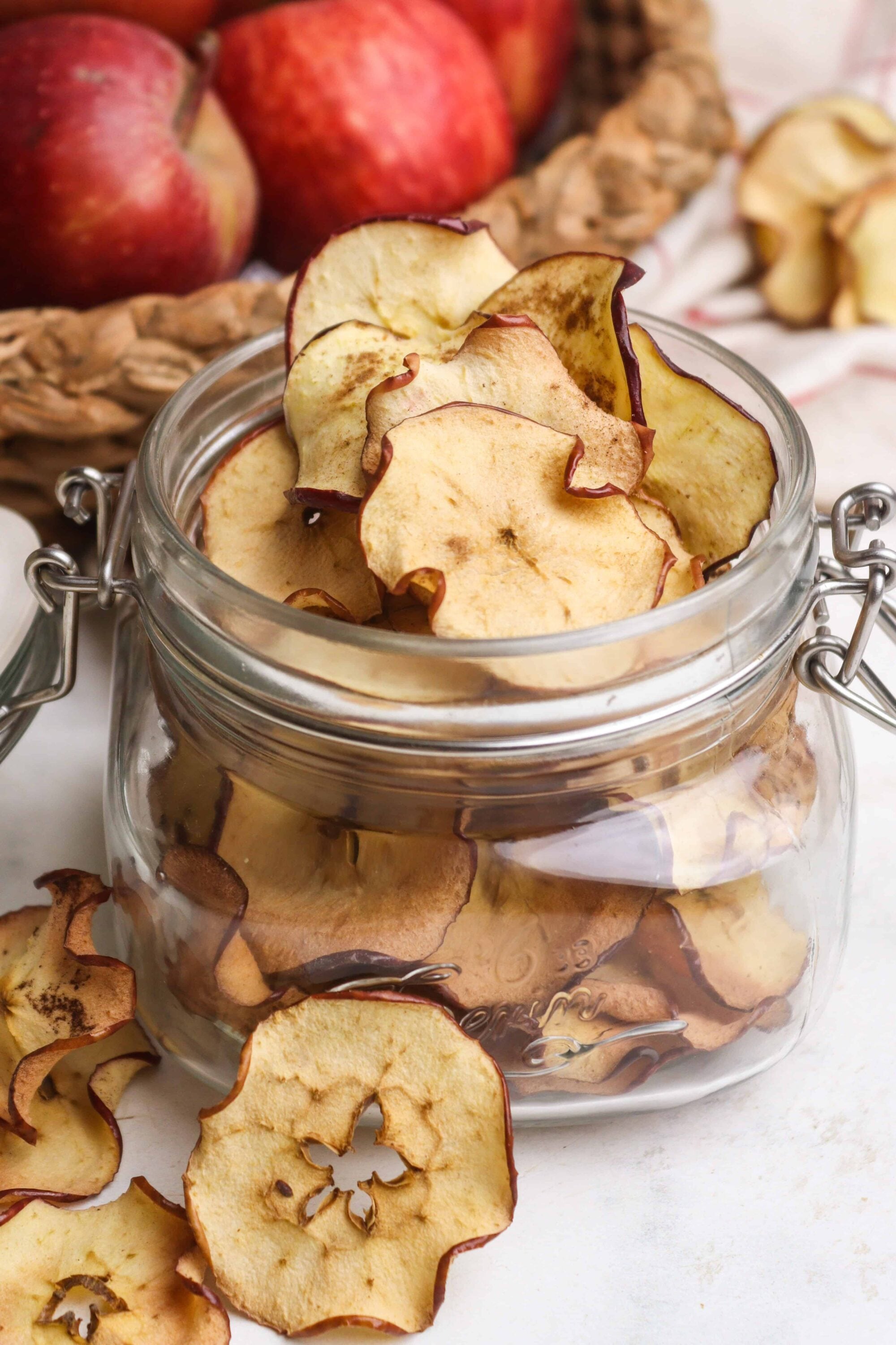 Dried apple slices in a glass jar with fresh apples and apple peels in background.