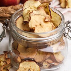 Dried apple slices in a glass jar with fresh apples and apple peels in background.