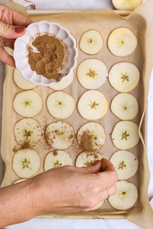 Sliced apples with cinnamon being prepared for baking, cinnamon powder in dish, apple rings on baking sheet.