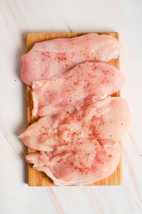 Fresh raw chicken breasts garnished with spices on a wooden cutting board.