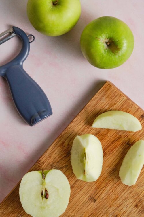 Fresh green apples with sliced halves on a wooden cutting board, apple peeler in background, perfect for healthy snack recipes.