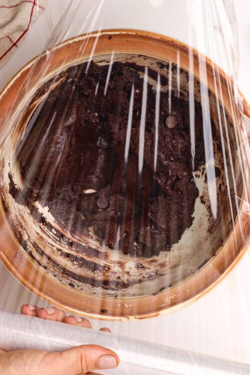 Fudgy chocolate cake baking in a copper bundt pan under plastic wrap.