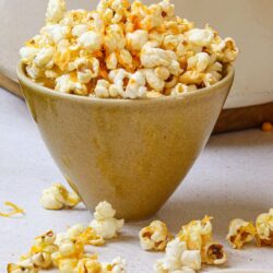 Buttered popcorn in a beige bowl with scattered popcorn and seasoning on light background.