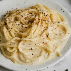Creamy alfredo pasta with black pepper and shaved parmesan cheese on a white speckled plate.