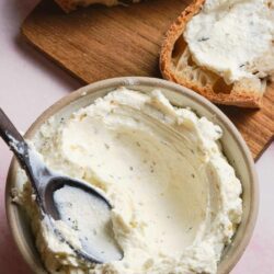 Creamy homemade butter in a bowl with bread slices with butter on a wooden cutting board.