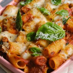 Baked pasta with melted cheese, fresh basil, and tomato sauce in a pink baking dish.