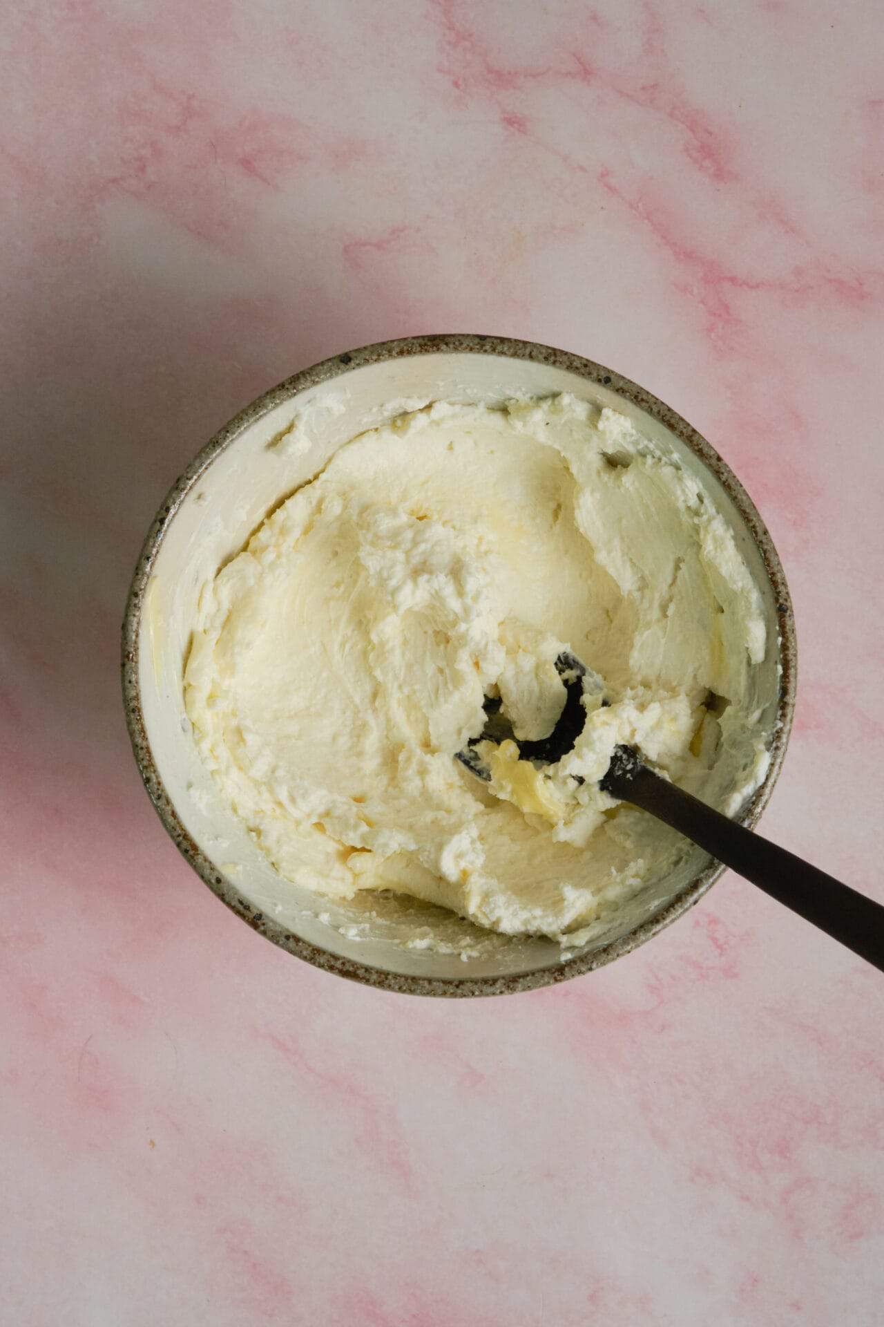 Creamy homemade butter in a bowl with a black spatula, on a pink textured background.