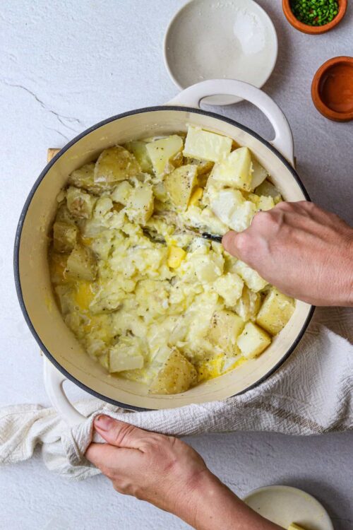 Creamy mashed potatoes in a dish with hands mixing and seasoning, hearty comfort food.