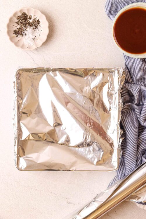 Baked Bree's oven with foil-covered baking dish on a light countertop, surrounding ingredients and cooking essentials.