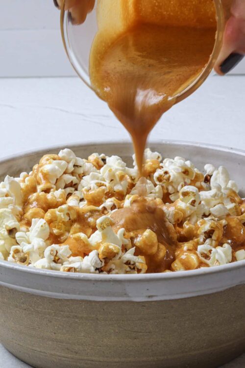 Rich caramel sauce being poured over fluffy popcorn in a white bowl, perfect for sweet snacks.