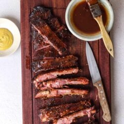 Tender smoked ribs with barbecue sauce on a wooden cutting board, surrounded by mustard, mustard, and barbecue seasoning.