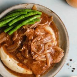 Savory pork chop smothered in onion gravy with green beans and mashed potatoes on a rustic plate.