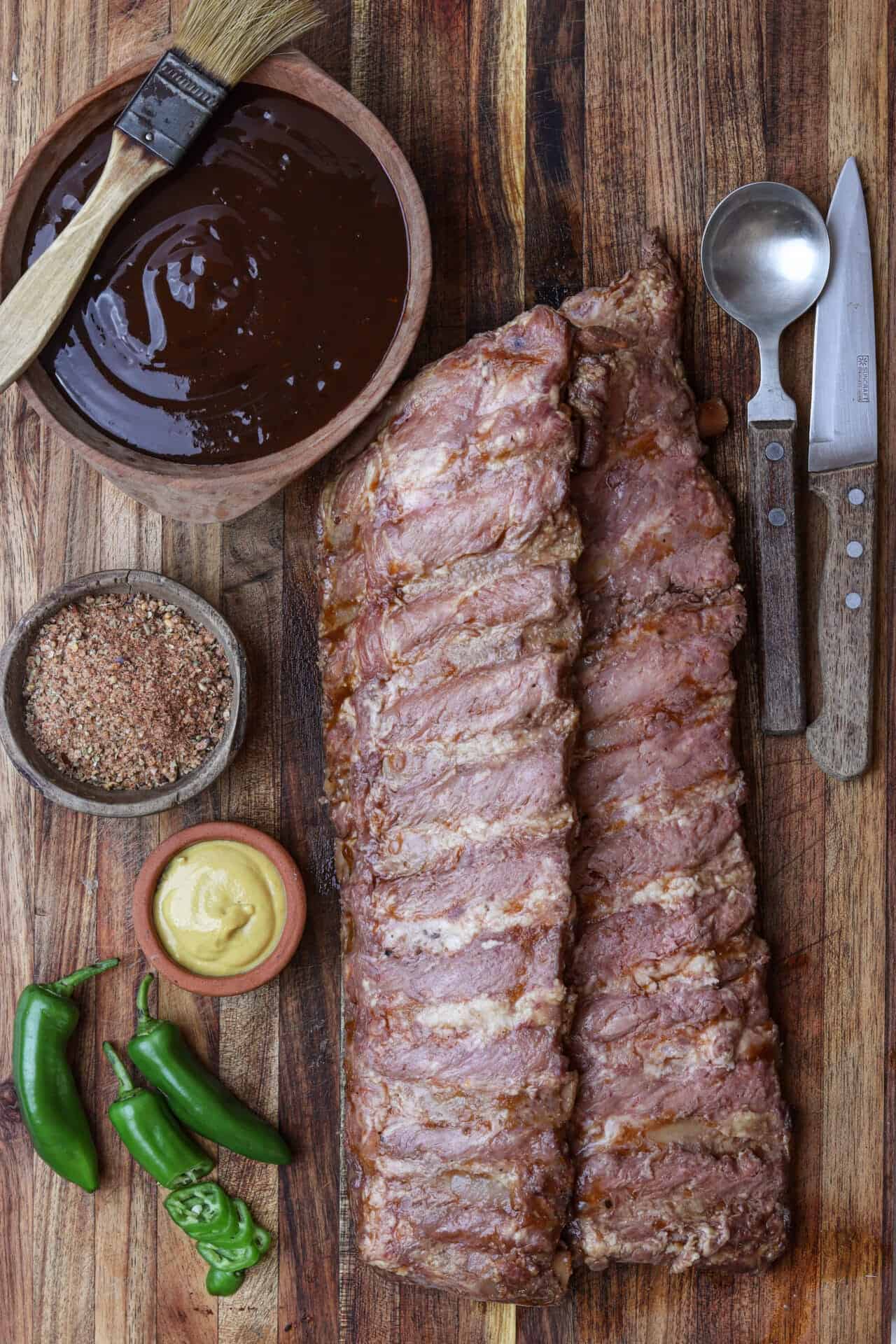 Juicy pork ribs with barbecue glaze on wooden cutting board, grill sauce, spices, mustard, peppers, and kitchen tools ready for cooking.