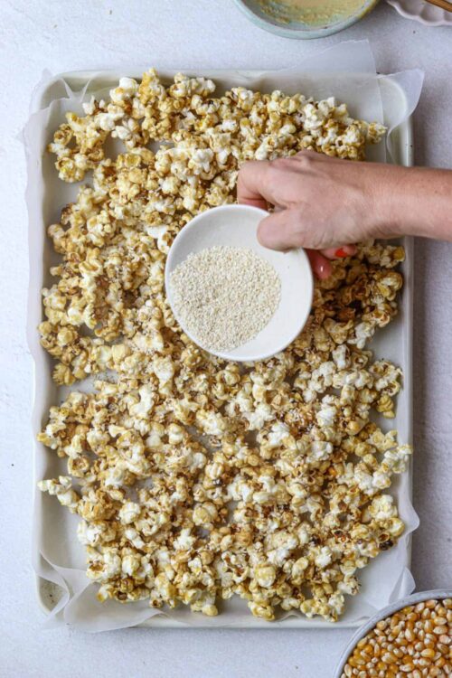 Light caramel popcorn being sprinkled with sesame seeds, prepared for baking.