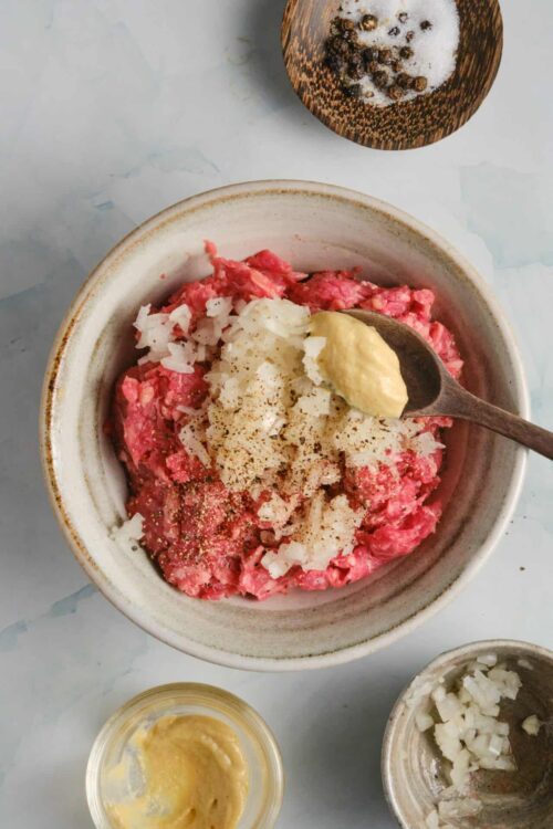 Finely chopped raw ground beef with onions, seasonings, and mustard in a bowl for burger patties.