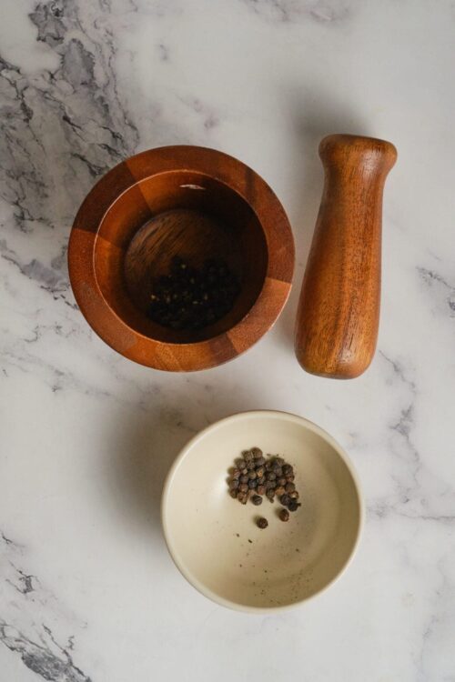 Whole black peppercorns in a beige bowl and a wooden pepper grinder on a white marble surface.