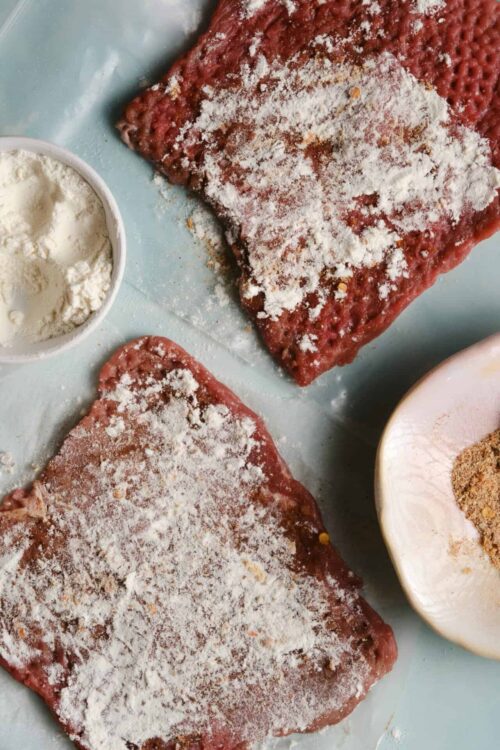 Raw seasoned beef steak with flour, salt, pepper, and spices on light blue surface.