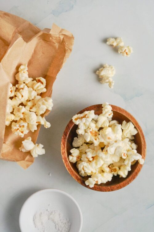 Fluffy popcorn in a brown paper bag, on a light marble surface with a small white bowl of salt.