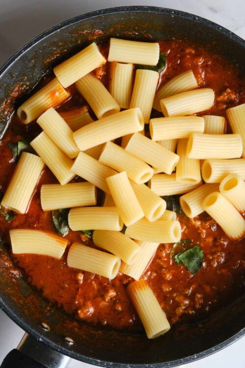 Canned pasta with tomato sauce and uncooked rigatoni pasta in a skillet.