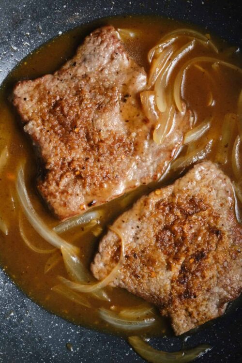 Juicy pork chops cooking in a skillet with onions and savory broth.