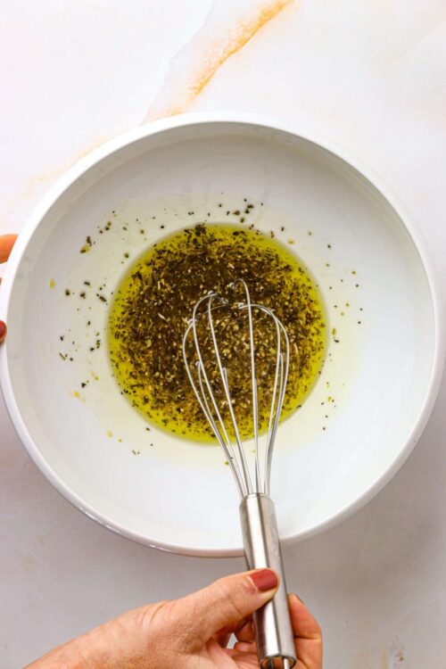 Olive oil and herb mixture in a white mixing bowl with a whisk.
