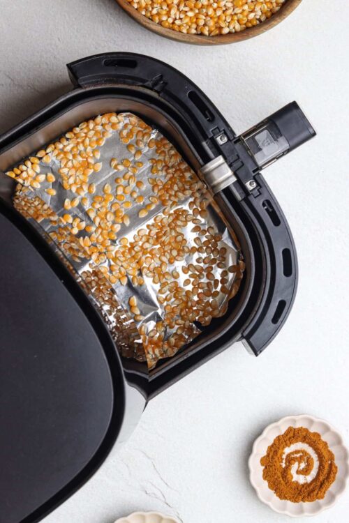 Unpopped popcorn kernels in an air fryer with a bowl of seasoning on the side.