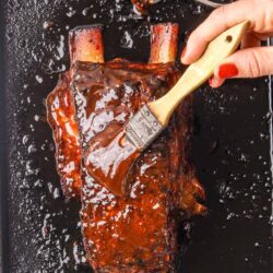 Glazed rack of ribs being coated with barbecue sauce on a baking sheet.