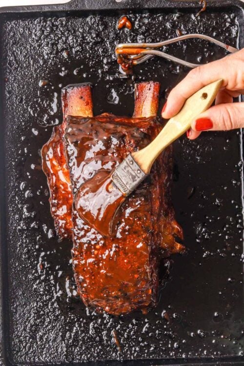 Glazed rack of ribs being coated with barbecue sauce on a baking sheet.