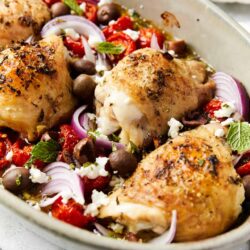 Roast chicken thighs with cherry tomatoes, olives, and herbs in a baking dish.