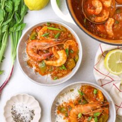 Fresh shrimp stew with vegetables served over rice, garnished with green onions, with fresh herbs and lemon on a white background.