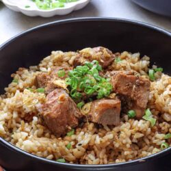 Savory braised beef short rib with rice in black bowl, garnished with green onions, on gray surface.
