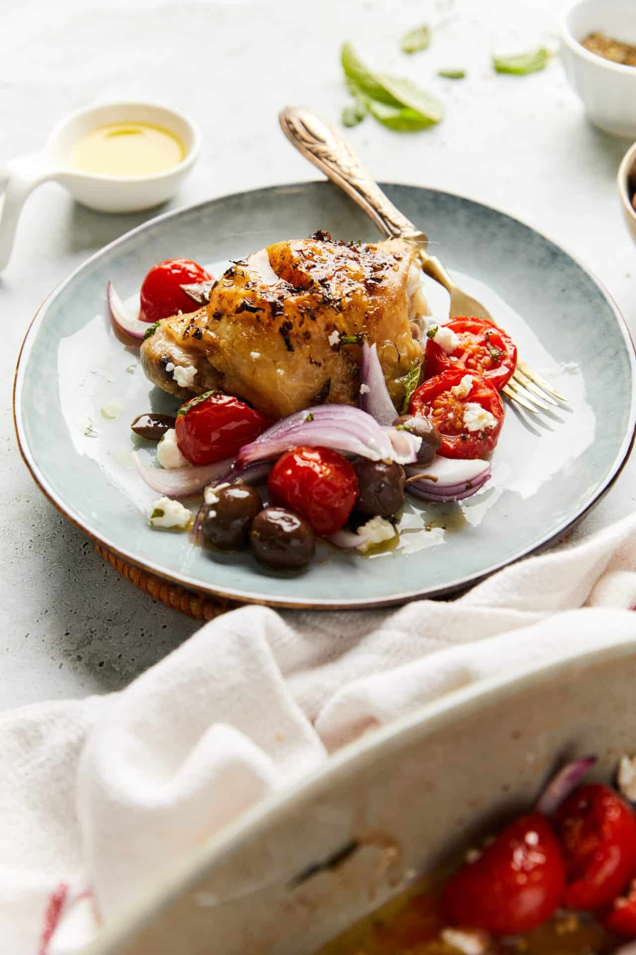 Roasted chicken with cherry tomatoes, red onions, and olives on a plate, side dishes and cut lime in background.