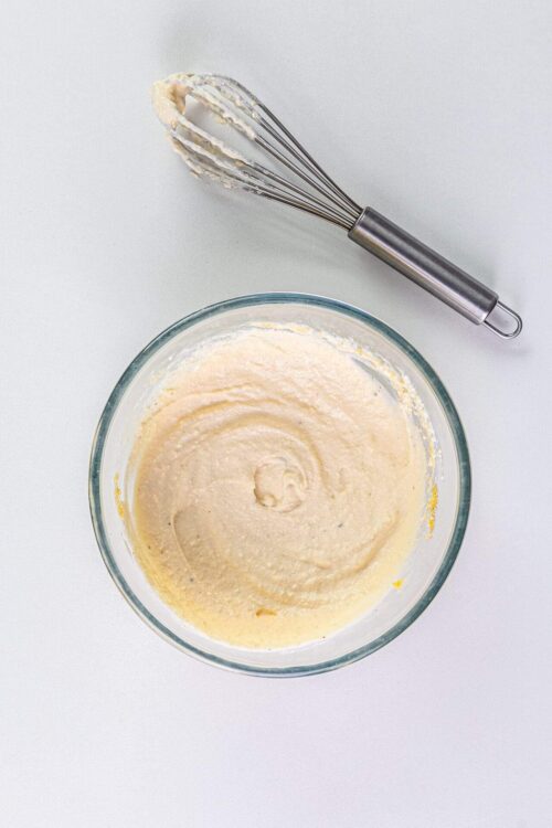 Beaten cake batter in glass mixing bowl with whisk on white surface.