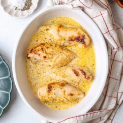 Juicy baked chicken breasts in creamy sauce with herbs on white baking dish.