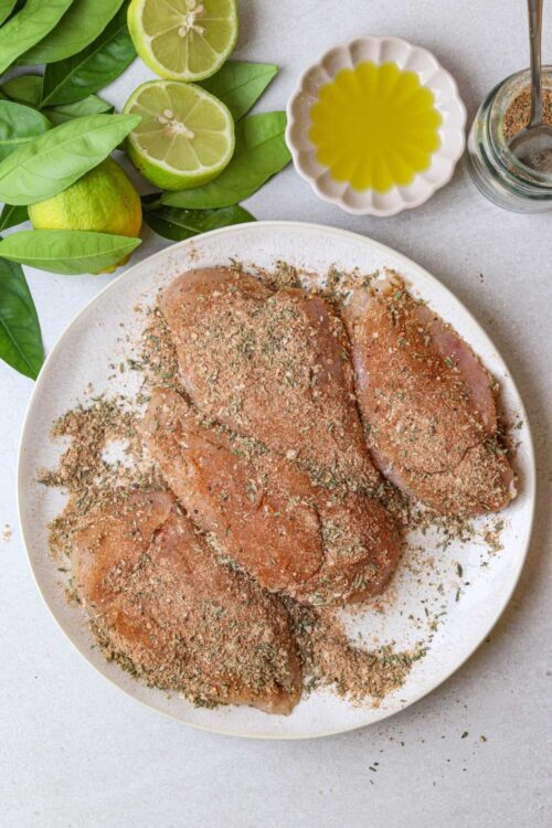 Baked chicken breasts seasoned with herbs on a white plate, with fresh limes, basil leaves, and olive oil nearby.