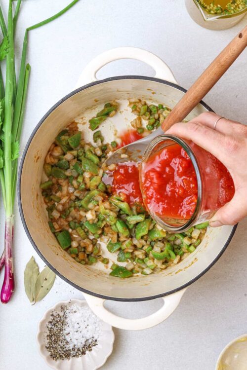 Chopped green vegetables being cooked in a pot with tomato sauce being poured in for a flavorful dish.